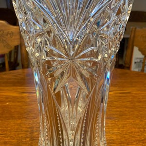 May include: A clear, cut-glass vase with a starburst design at the base and a flared top. The vase has vertical and geometric patterns, creating a textured appearance. The vase is sitting on a wooden table.