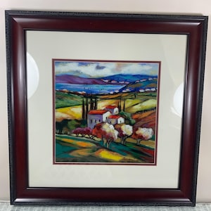 May include: Framed artwork depicting a colorful landscape with houses, trees, and a body of water. The painting features a vibrant palette of blues, greens, yellows, and reds. The frame is a dark, ornate wood.
