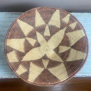 May include: Round woven basket with a starburst design. The basket features a central star pattern in light beige, surrounded by alternating brown and beige triangular shapes. The rim is a darker brown.
