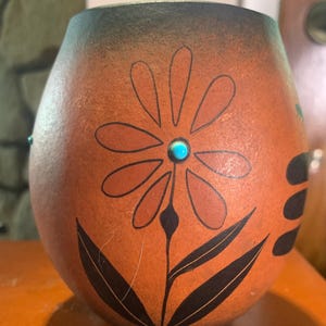 May include: A hand-painted, gourd-shaped vase with a floral design. The vase is primarily orange with a black and turquoise flower design. The top of the vase is dark brown.