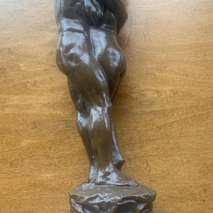 May include: Bronze sculpture depicting two figures embracing. The figures are standing on a textured rock base atop a rectangular block. The sculpture has a dark brown patina and is set against a wooden background.