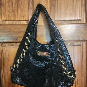 May include: A black hobo bag with a snakeskin-embossed texture, featuring thick gold-tone chain links on both sides and a central gold-tone metal plaque engraved with the text MICHAEL KORS EST. 1981. The bag has a single shoulder strap.
