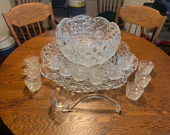 May include: A vintage clear glass punch bowl set with a large bowl, a serving tray, a ladle, and several small cups. The set is made of cut glass with a detailed pattern, and is displayed on a round wooden table.