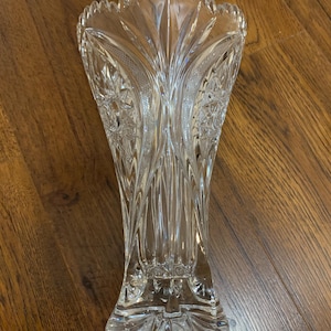 May include: A clear, cut-glass vase with a flared top and a detailed, textured pattern. The vase features a wide base and a series of vertical lines and starburst designs. The rim has a scalloped edge.