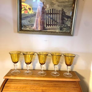 May include: Five amber-colored wine glasses with a textured surface sit on a wooden surface. The glasses have a clear stem and a round base.