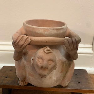 May include: A terracotta-colored ceramic planter with a unique design. The planter features a sculpted figure with arms holding a rim, and a face on the front. The planter is sitting on a small wooden shelf.