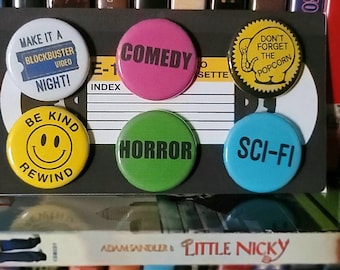 Könnte beinhalten: Ein Satz von sieben runden Knöpfen mit verschiedenen Filmgenre-Themen. Die Knöpfe sind gelb, blau, grün, rosa und rot mit schwarzem Text. Der Text auf den Knöpfen lautet "Make it a Blockbuster Video Night!" "Be Kind Rewind" "Comedy" "Horror" "Sci-Fi" "Don't Forget the Popcorn"