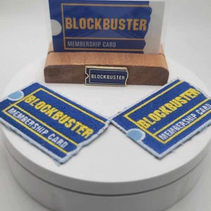 May include: A selection of Blockbuster-themed merchandise. Features a membership card, two embroidered patches, and a pin. The patches and card display the Blockbuster logo in blue and yellow. The pin is a small version of the logo.