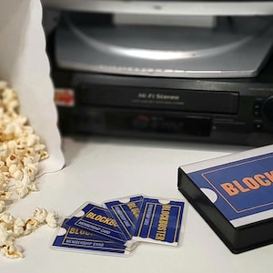 May include: A black VCR player, a white paper bag filled with popcorn, and several blue and orange Blockbuster membership cards arranged next to a rectangular box featuring the Blockbuster logo in orange text on a blue background.