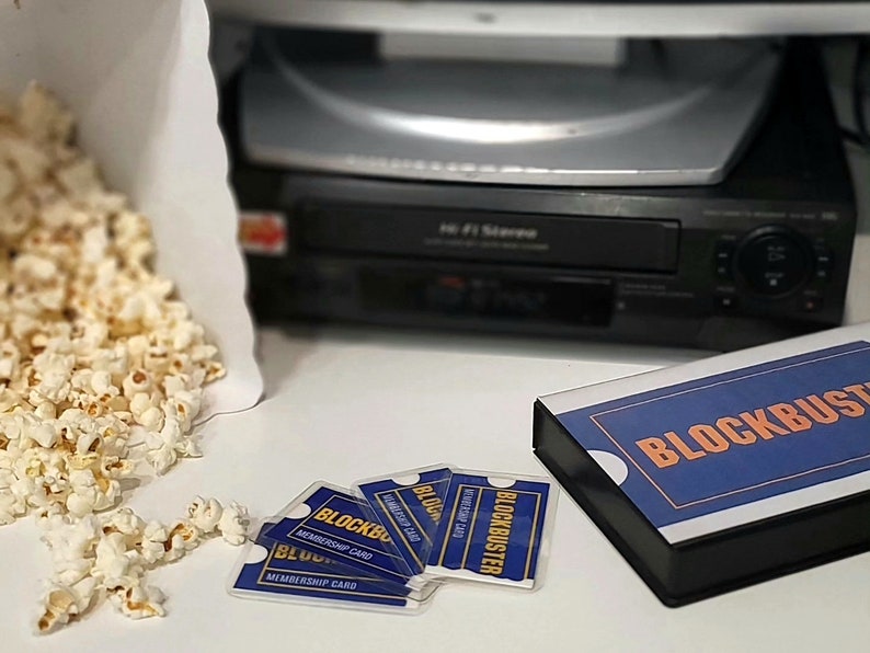 May include: A black VCR player, a white paper bag filled with popcorn, and several blue and orange Blockbuster membership cards arranged next to a rectangular box featuring the Blockbuster logo in orange text on a blue background.