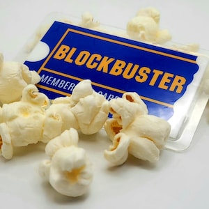 May include: A rectangular blue and yellow Blockbuster membership card encased in a clear plastic sleeve, partially covered by several pieces of white, popped popcorn kernels scattered across the surface of the card.