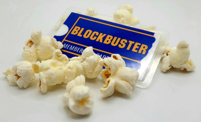 May include: A rectangular blue and yellow Blockbuster membership card encased in a clear plastic sleeve, partially covered by several pieces of white, popped popcorn kernels scattered across the surface of the card.
