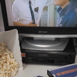 May include: A television screen displaying a scene from a movie, a black VCR, a white paper container filled with popcorn, and several blue and white Blockbuster membership cards next to a rectangular box labeled Blockbuster in blue and orange.