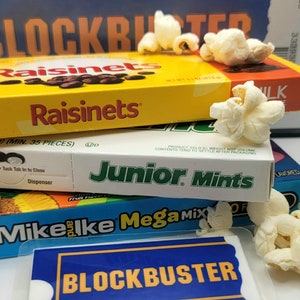 May include: A stack of candy boxes including Raisinets, Junior Mints, and Mike and Ike Mega Mix, topped with popcorn. In front lies a blue and yellow Blockbuster membership card in a clear plastic sleeve, with a Blockbuster box in the background.