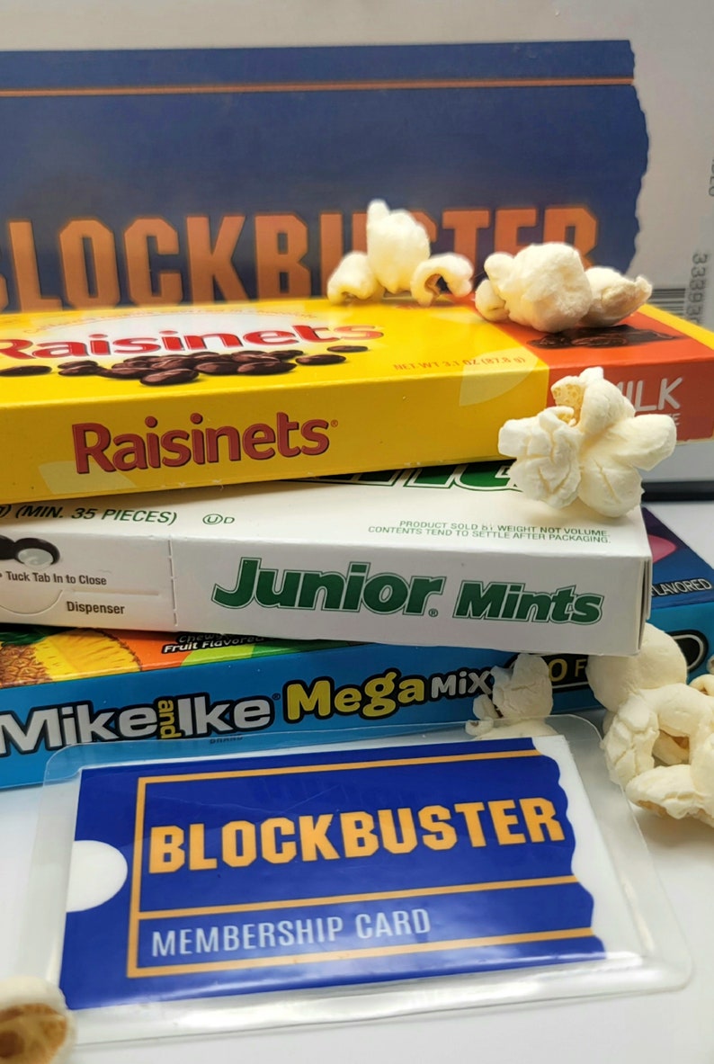 May include: A stack of candy boxes including Raisinets, Junior Mints, and Mike and Ike Mega Mix, topped with popcorn. In front lies a blue and yellow Blockbuster membership card in a clear plastic sleeve, with a Blockbuster box in the background.