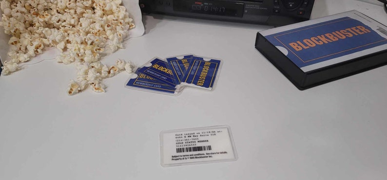 May include: A collection of blue and yellow Blockbuster membership cards with white text, a Blockbuster video case, and a pile of popcorn. One card is displayed face-up showing text including Gold Status Member, a barcode, and store details.