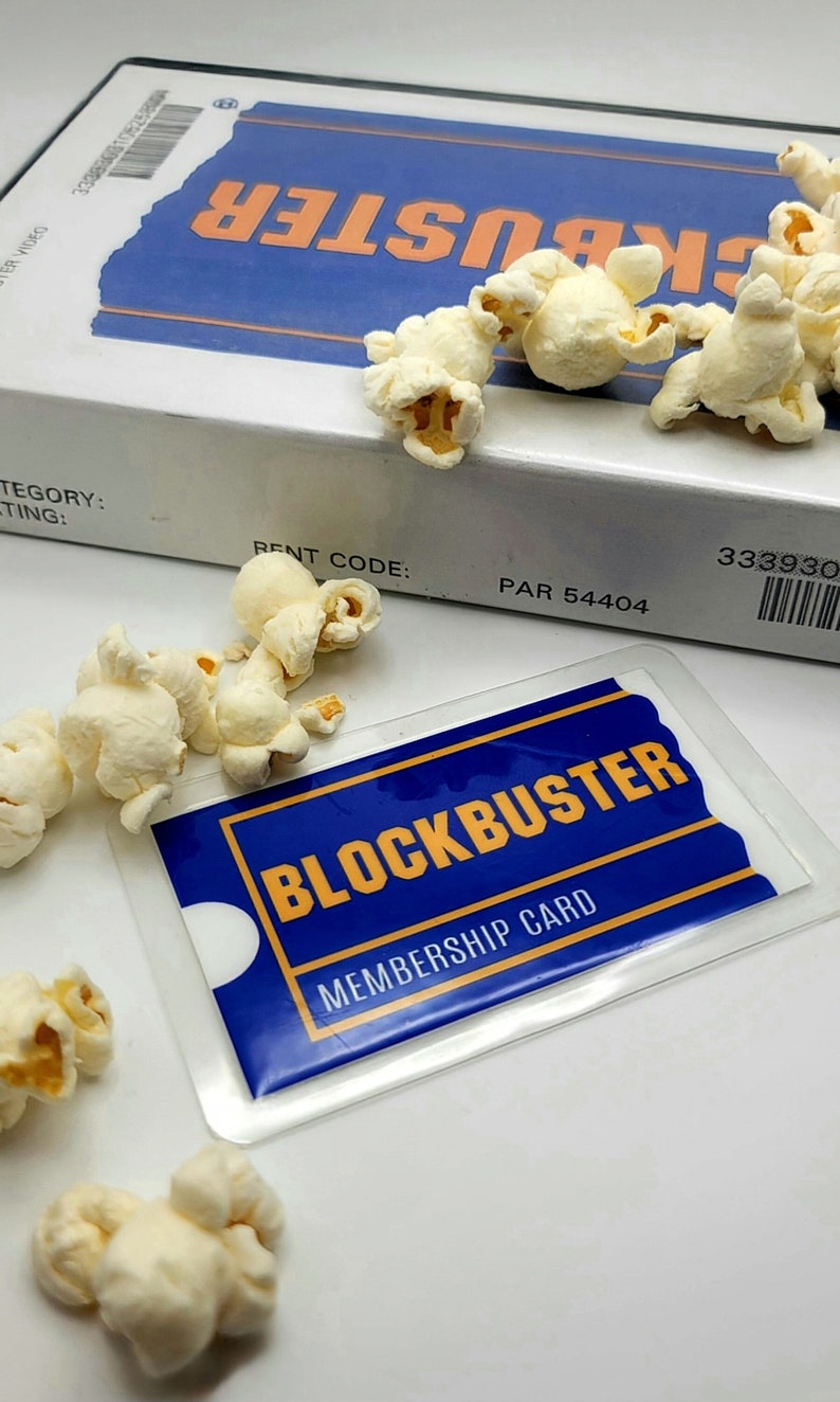 May include: A Blockbuster VHS case and a Blockbuster membership card in a clear plastic sleeve, surrounded by scattered pieces of white popcorn. The VHS case features the blue and orange logo, while the card displays the text Blockbuster Membership Card.