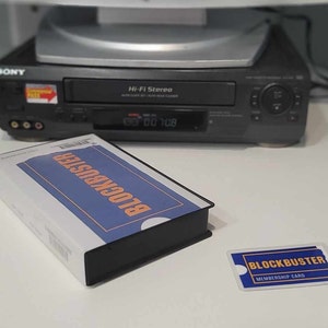 May include: A Blockbuster VHS tape in a white and blue sleeve with orange text, a matching Blockbuster membership card, and a black Sony Hi-Fi Stereo VCR in the background. The VCR features a digital display and a small red and yellow sticker.