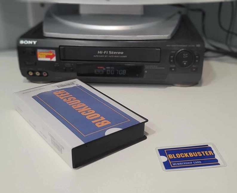 May include: A Blockbuster VHS tape in a white and blue sleeve with orange text, a matching Blockbuster membership card, and a black Sony Hi-Fi Stereo VCR in the background. The VCR features a digital display and a small red and yellow sticker.