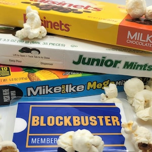 May include: A stack of candy boxes including Raisinets, Junior Mints, and Mike and Ike Mega Mix, topped with scattered popcorn. In the foreground, a blue and yellow Blockbuster membership card is visible, also surrounded by pieces of popcorn.