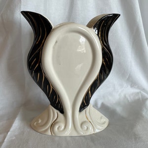 May include: A white ceramic vase with black and gold accents. The vase has a unique shape with two wings on either side. The wings are decorated with black and gold stripes.