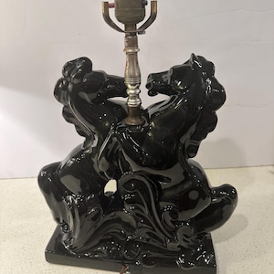 Op de afbeelding: Zwarte keramische tafellamp met twee steigerende paarden. De lampvoet is glanzend zwart met ingewikkelde details. De lamp heeft een standaard lamphouder en een bruin netsnoer.