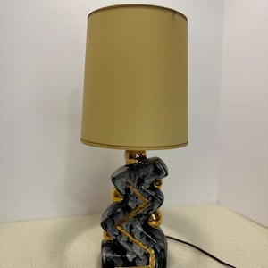 Puede incluir: Lámpara de mesa de cerámica con pantalla beige. La base es negra con detalles grises y dorados. La lámpara tiene un diseño abstracto único y un cable de alimentación marrón.