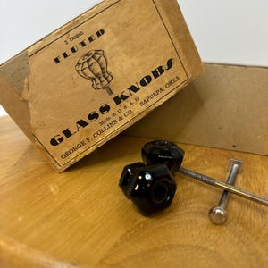 May include: Two black fluted glass knobs with a box labeled "GLASS KNOBS" and "Made in U.S.A." The knobs are next to a screw and a wooden surface. The box reads "2 Dozen FLUTED".