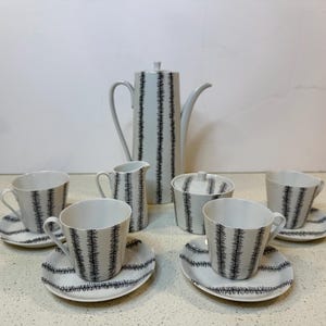 May include: A white porcelain tea set with a black vertical line pattern. The set includes a coffee pot, creamer, sugar bowl, and six cups with saucers. The set is designed for serving tea or coffee.