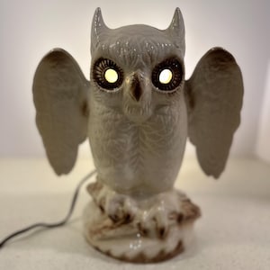 May include: A ceramic owl figurine with glowing yellow eyes and outstretched wings. The owl is light gray with brown accents and sits on a decorative base. A black cord extends from the base.