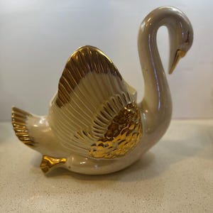 Puede incluir: Un macetero de cerámica blanca en forma de cisne con detalles dorados. Las alas del cisne están extendidas y el macetero tiene un borde dorado.
