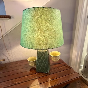 Może przedstawiać: Lampa stołowa z teksturowanym zielonym abażurem i ciemnozieloną ceramiczną podstawą z reliefem w kształcie rombów. Podstawa posiada dwie małe, jasne żółte okrągłe doniczki lub miseczki przymocowane po bokach kolumny centralnej.