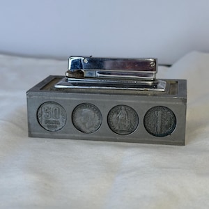 May include: A silver-colored lighter with a rectangular base. The base features four coin insets. The lighter is positioned on top of the base. The coins are visible in the base.