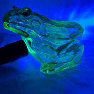 May include: A translucent, lime-green glass frog figurine illuminated by a blacklight, causing it to glow intensely. The frog is depicted in a seated position with molded details representing its eyes, limbs, and textured skin, captured under UV light.