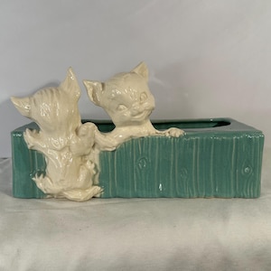 May include: A vintage ceramic planter in a rectangular shape, featuring two cream-colored cat figurines peeking over a teal-colored wooden fence design. The planter is a decorative item.