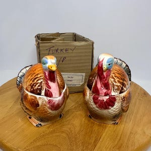 May include: Two ceramic turkey figurines with open tops, featuring brown, orange, and red plumage. The turkeys have blue and yellow heads and are on a wooden surface. A cardboard box with the text "TURKEY" is in the background.