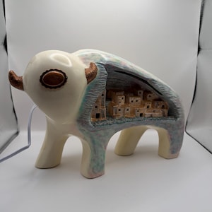 May include: A ceramic bison-shaped lamp with a glossy cream-colored exterior and copper-toned horns. The side of the bison features a hollowed-out cutout revealing a detailed miniature desert village scene with tan buildings and ladders inside.