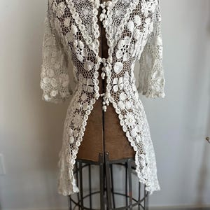 May include: A white crocheted top with a button-up front and a delicate floral pattern. The top has a long, flowing silhouette and is perfect for layering.