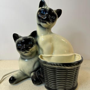 Op de afbeelding: Vintage Siamese kattenbeeldje met een mand. De keramische katten zitten, met zwart-crème kleuring en blauwe ogen. De mand is donkergrijs. Een netsnoer loopt vanaf de basis.