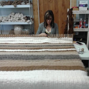 May include: A woman weaving a rug on a loom. The rug features horizontal stripes in white, brown, and grey. Shelves in the background hold spools of yarn. The woman wears glasses and a grey cardigan.