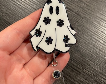May include: A white ghost with black glitter accents and a black glitter face. The ghost is attached to a clear plastic retractable badge holder with a black glitter button.