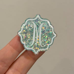 May include: A decorative, glittery monogram initial "M" in white, set against a teal background. The monogram is encased in a scalloped, light teal frame. The background is filled with iridescent glitter.