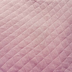 Peut inclure: Gros plan sur un tissu matelass&eacute; de couleur rose p&acirc;le et douce. Le tissu pr&eacute;sente un motif en losanges cr&eacute;&eacute; par des lignes cousues, ajoutant de la texture et de l'int&eacute;r&ecirc;t visuel. Le mat&eacute;riau semble doux et moelleux.