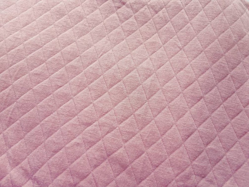 Peut inclure: Gros plan sur un tissu matelass&eacute; de couleur rose p&acirc;le et douce. Le tissu pr&eacute;sente un motif en losanges cr&eacute;&eacute; par des lignes cousues, ajoutant de la texture et de l'int&eacute;r&ecirc;t visuel. Le mat&eacute;riau semble doux et moelleux.