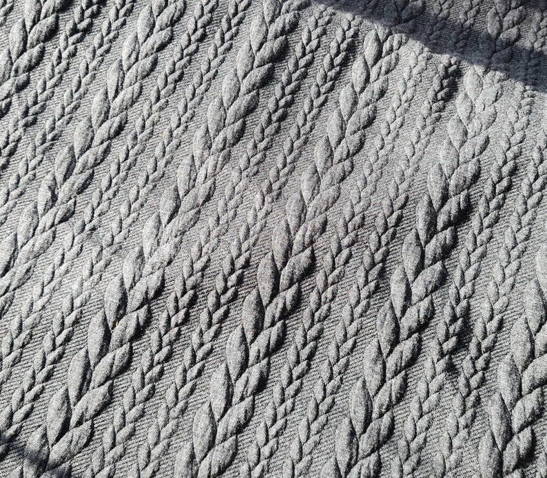 Peut inclure: Gros plan sur un tissu gris avec un motif r&eacute;p&eacute;t&eacute; de motifs en relief en forme de feuilles. La texture est d&eacute;taill&eacute;e, avec les feuilles dispos&eacute;es en diagonale. Le tissu semble doux et textur&eacute;.