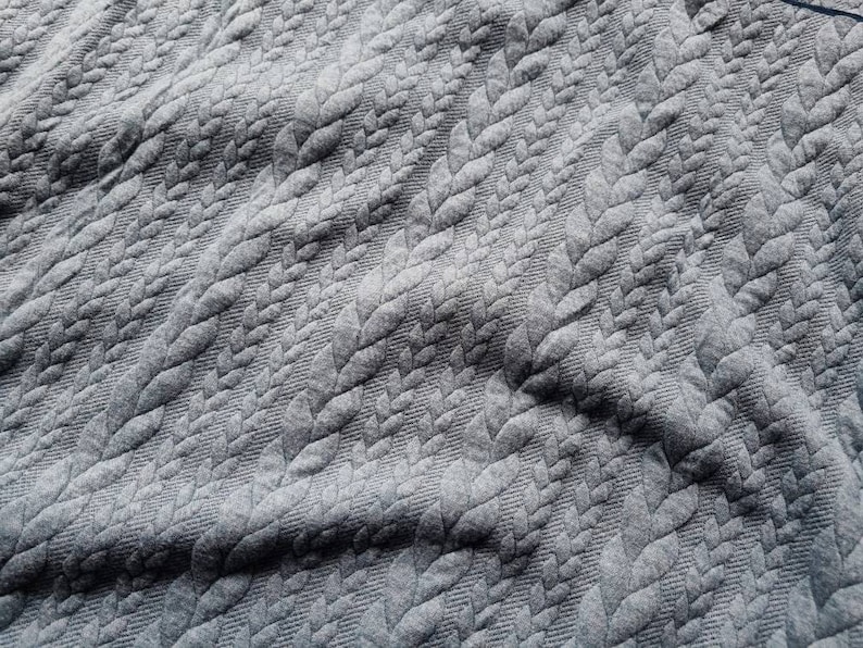 Peut inclure: Gros plan sur un tissu gris avec un motif de feuilles en relief. La texture cr&eacute;e un motif r&eacute;p&eacute;titif sur le mat&eacute;riau. Le tissu semble doux et souple, avec des plis et des ombres d&eacute;licats.