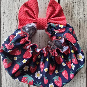 Könnte beinhalten: Ein marineblauer Stoff mit Erdbeerdruck und weißen Blumen ist gerafft und gerüscht, mit roter Bordüre. Eine große rote Schleife mit Polka-Dots ist am Stoff befestigt.