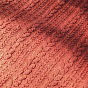 Peut inclure: Gros plan sur un tissu textur&eacute; de couleur corail. Le tissu pr&eacute;sente un motif r&eacute;p&eacute;titif de feuilles stylis&eacute;es dispos&eacute;es en diagonale. Les feuilles ont un aspect matelass&eacute; en relief, cr&eacute;ant une surface tactile. L'&eacute;clairage met en valeur la texture et la profondeur du motif.