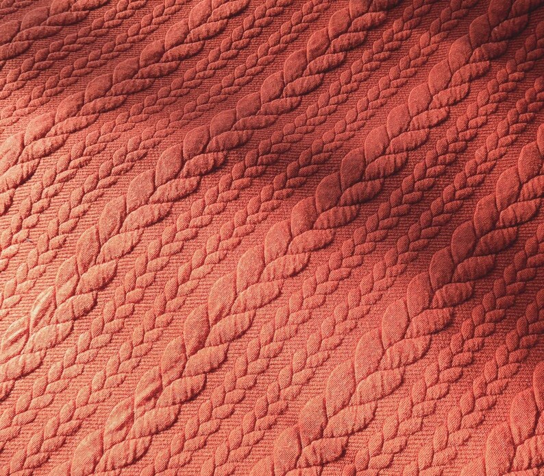 Peut inclure: Gros plan sur un tissu textur&eacute; de couleur corail. Le tissu pr&eacute;sente un motif r&eacute;p&eacute;titif de feuilles stylis&eacute;es dispos&eacute;es en diagonale. Les feuilles ont un aspect matelass&eacute; en relief, cr&eacute;ant une surface tactile. L'&eacute;clairage met en valeur la texture et la profondeur du motif.