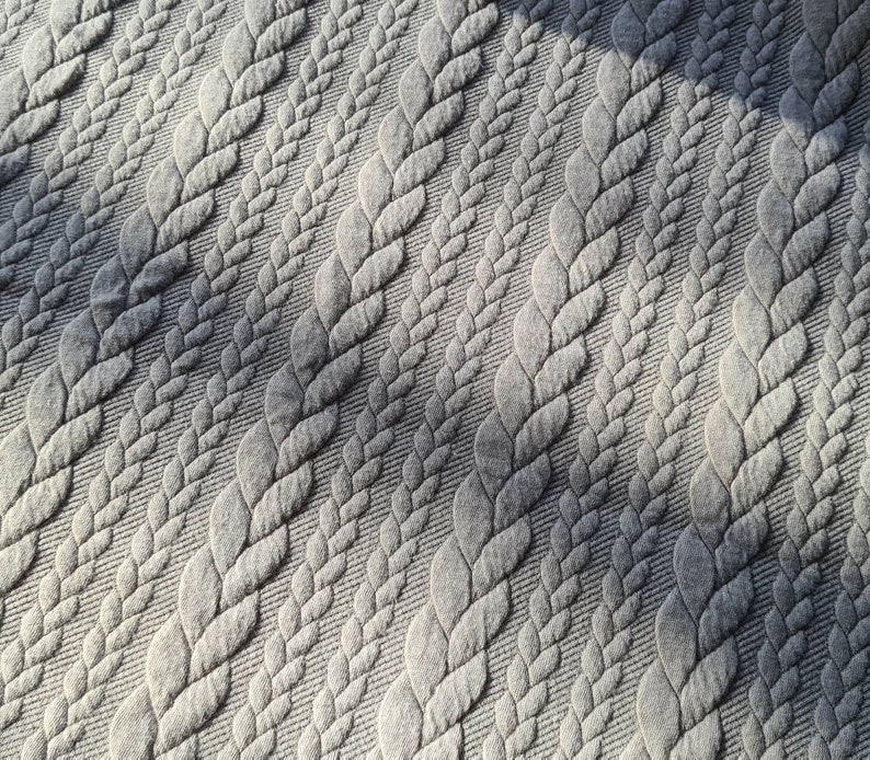 Peut inclure: Gros plan sur un tissu gris avec un motif r&eacute;p&eacute;t&eacute; de motifs tress&eacute;s en relief. La texture cr&eacute;e une sensation de profondeur et de dimension, les ombres accentuant l'aspect tiss&eacute;. La couleur du tissu est un gris neutre.